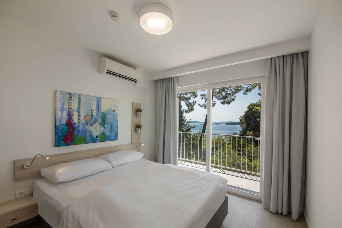 A modern bedroom with a large bed, a painting on the wall, and a balcony with a view of the sea.