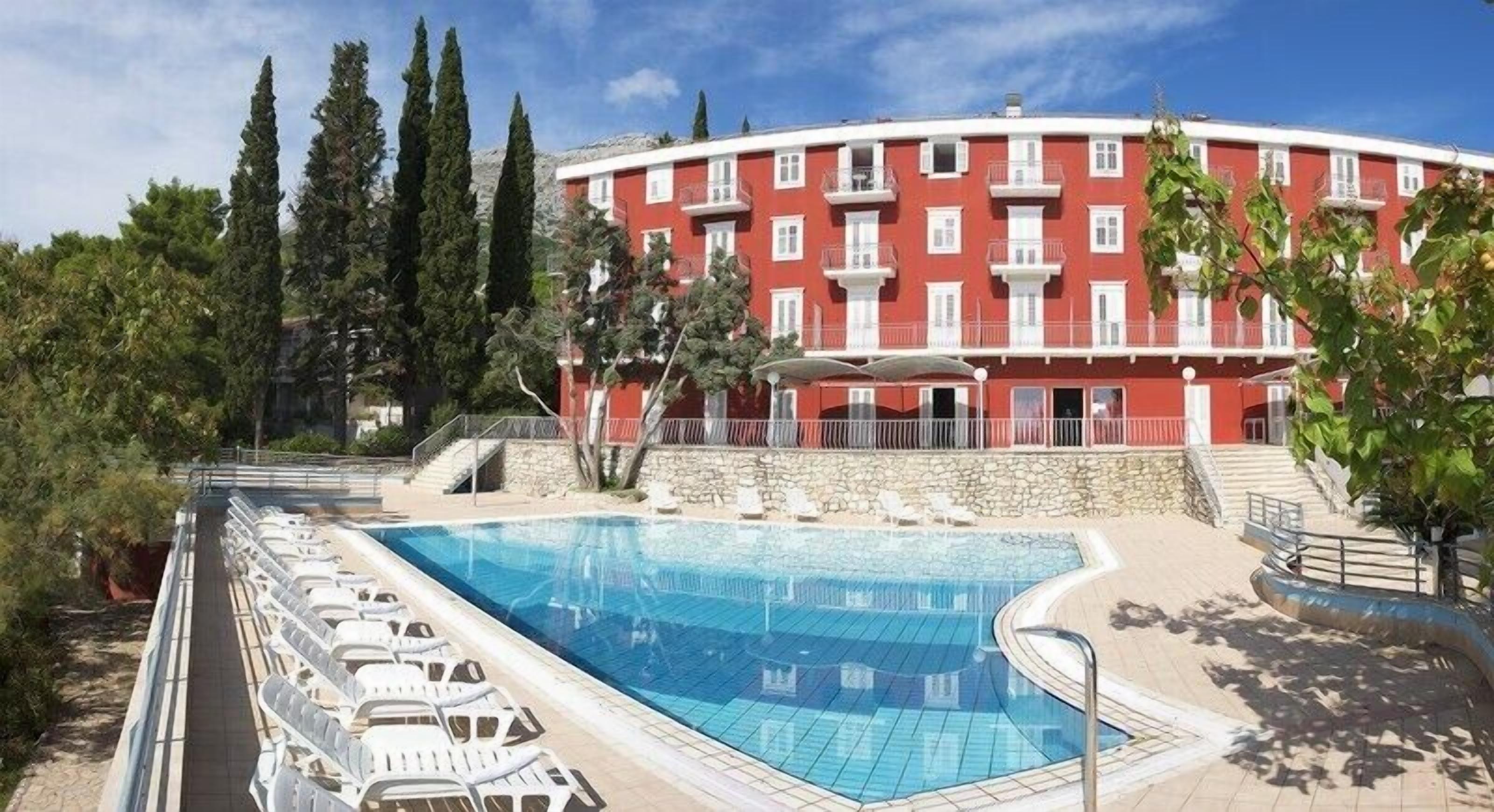 A swimming pool with lounge chairs, a red building with balconies, and trees in the background.