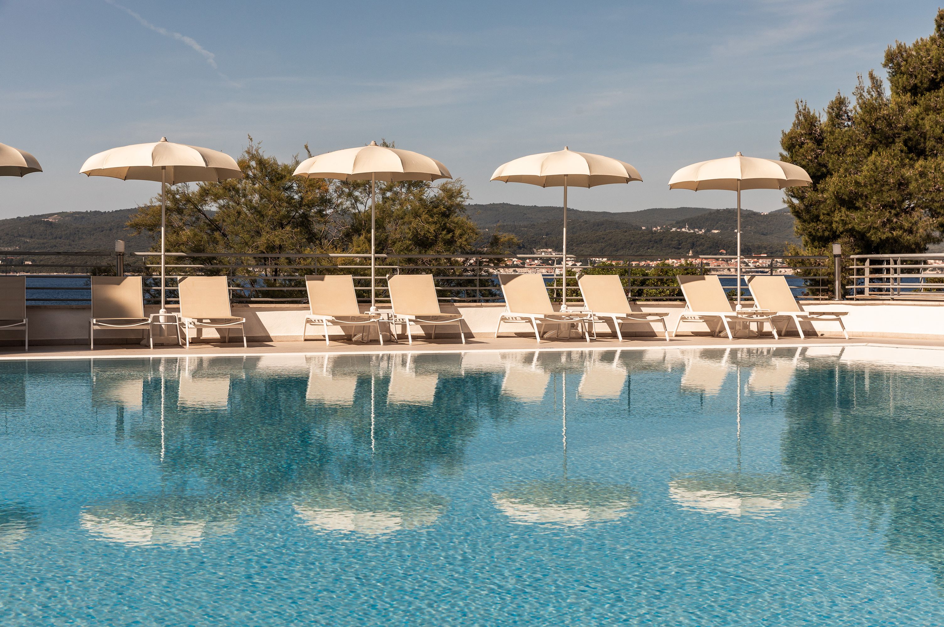A pool with clear blue water, surrounded by lounge chairs and umbrellas, with a view of hills in the background.