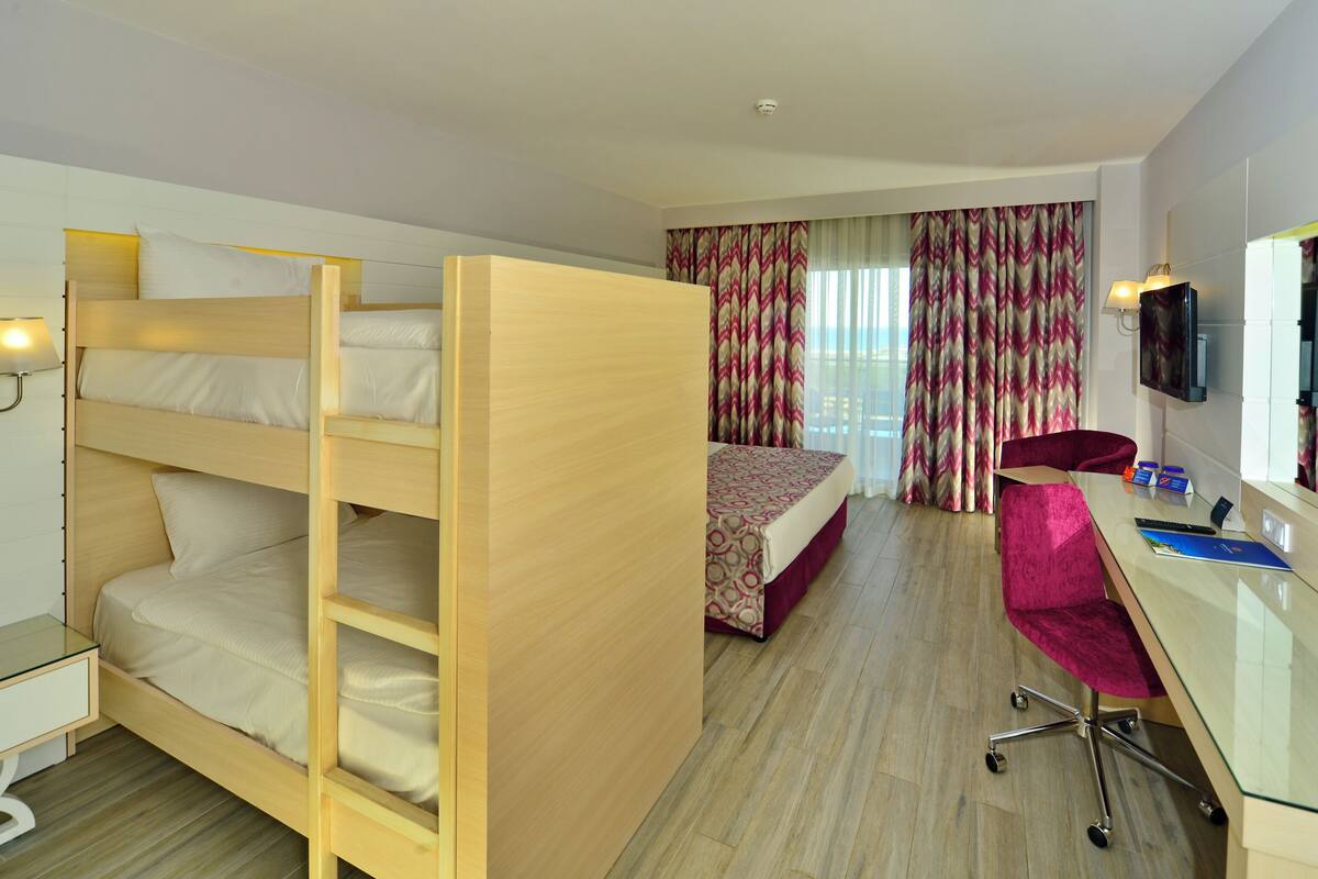 A hotel room with a bunk bed, a desk with a chair, a television, and a window with curtains.