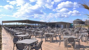 Un café en plein air avec de nombreuses tables et chaises, un espace de repos couvert et une vue sur la mer et les bâtiments en arrière-plan.