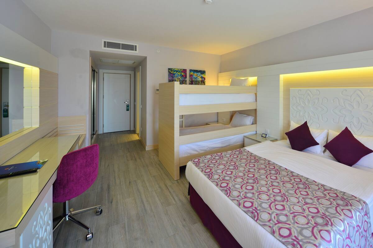 A hotel room with a bunk bed, a desk with a chair, a bedside table with a lamp, and a phone.