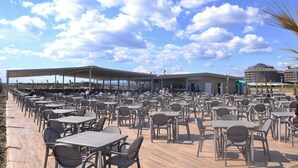 Een terras met veel tafels en stoelen, een overdekt zitgedeelte en uitzicht op een gebouw op de achtergrond.