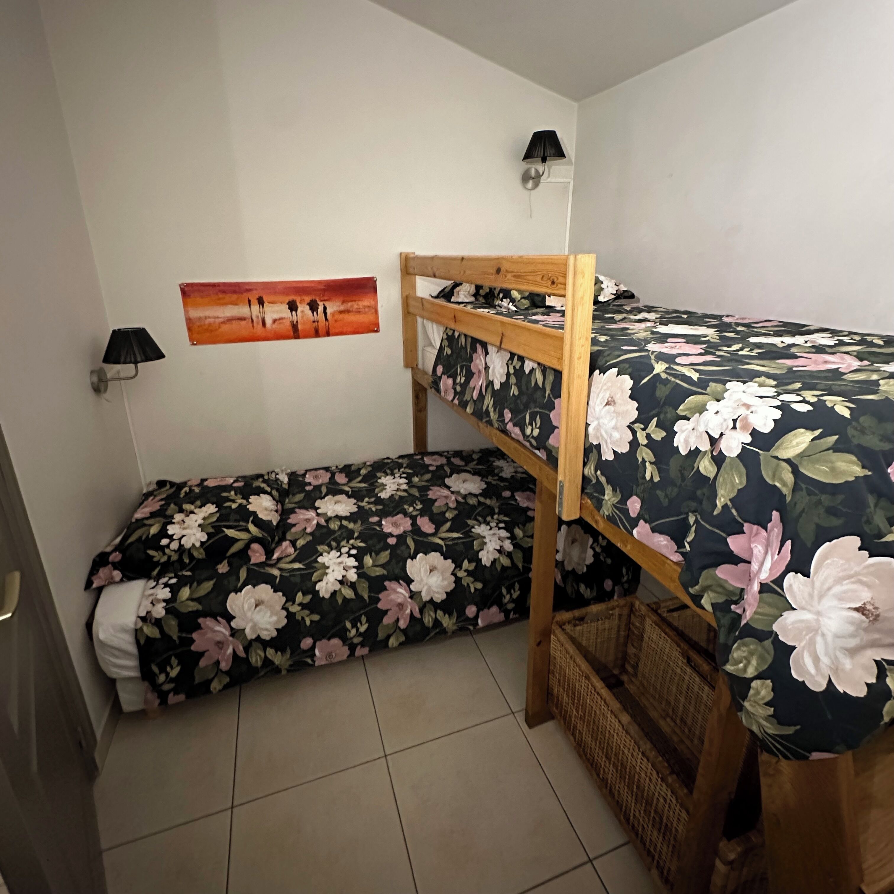 En våningssäng med blommigt sänglinne, en vägglampa och en tavla.