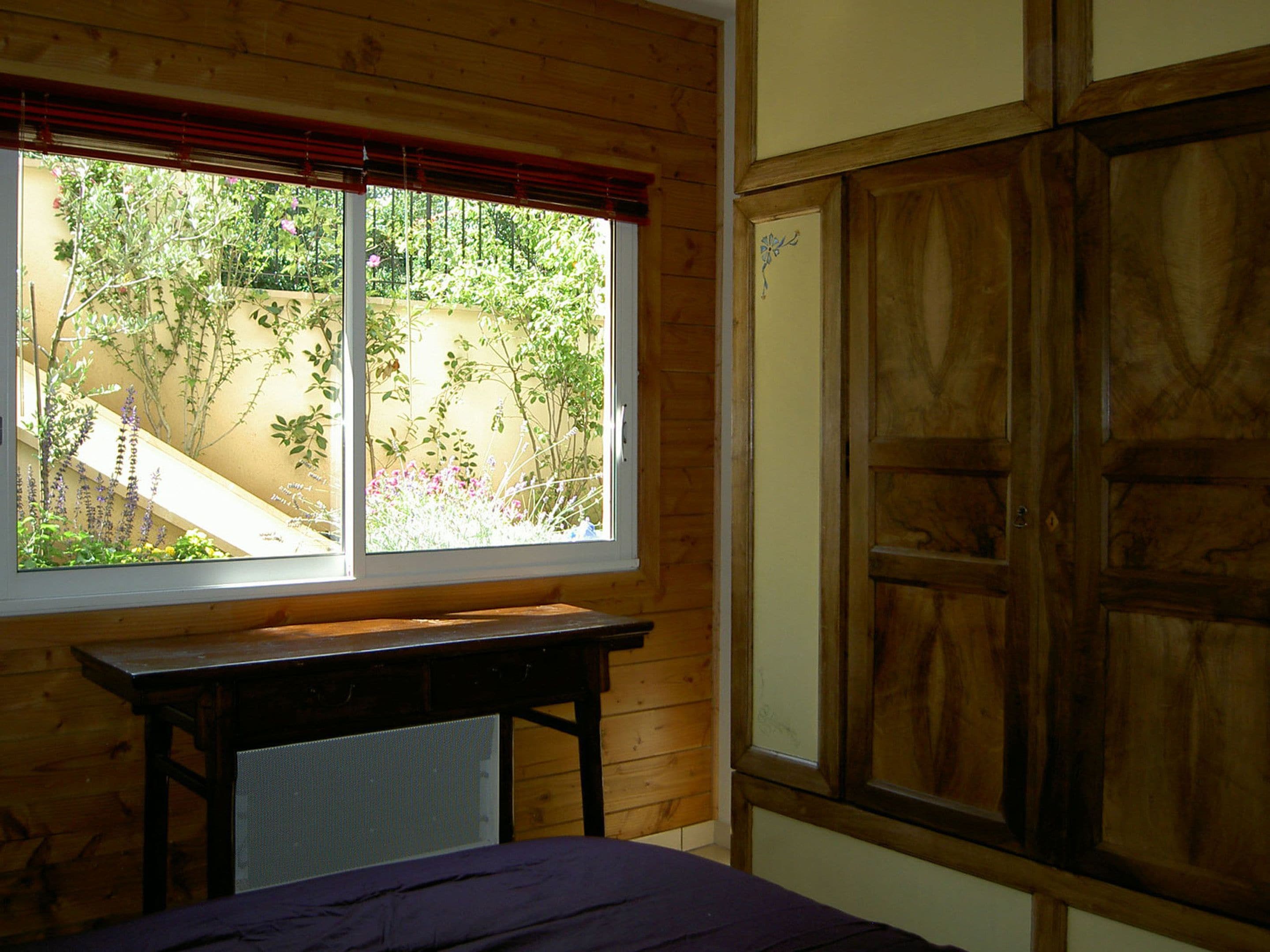 Een kamer met een houten bureau, een bed en een raam met uitzicht op een tuin.