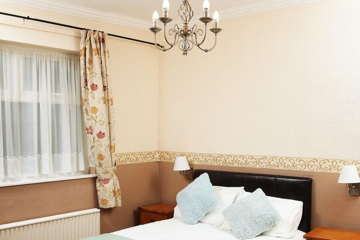 A neatly made bed with a red frame, a chandelier, a nightstand with a lamp, and a window with curtains.