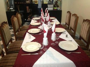 A dining table set with white plates, red napkins, and clear wine glasses, surrounded by wooden chairs. - Frantz Fanon (Setif)