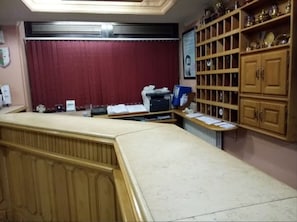 A reception area with a counter, a red curtain, a wooden cabinet with glass doors, and a framed portrait on the wall. - Frantz Fanon (Setif)