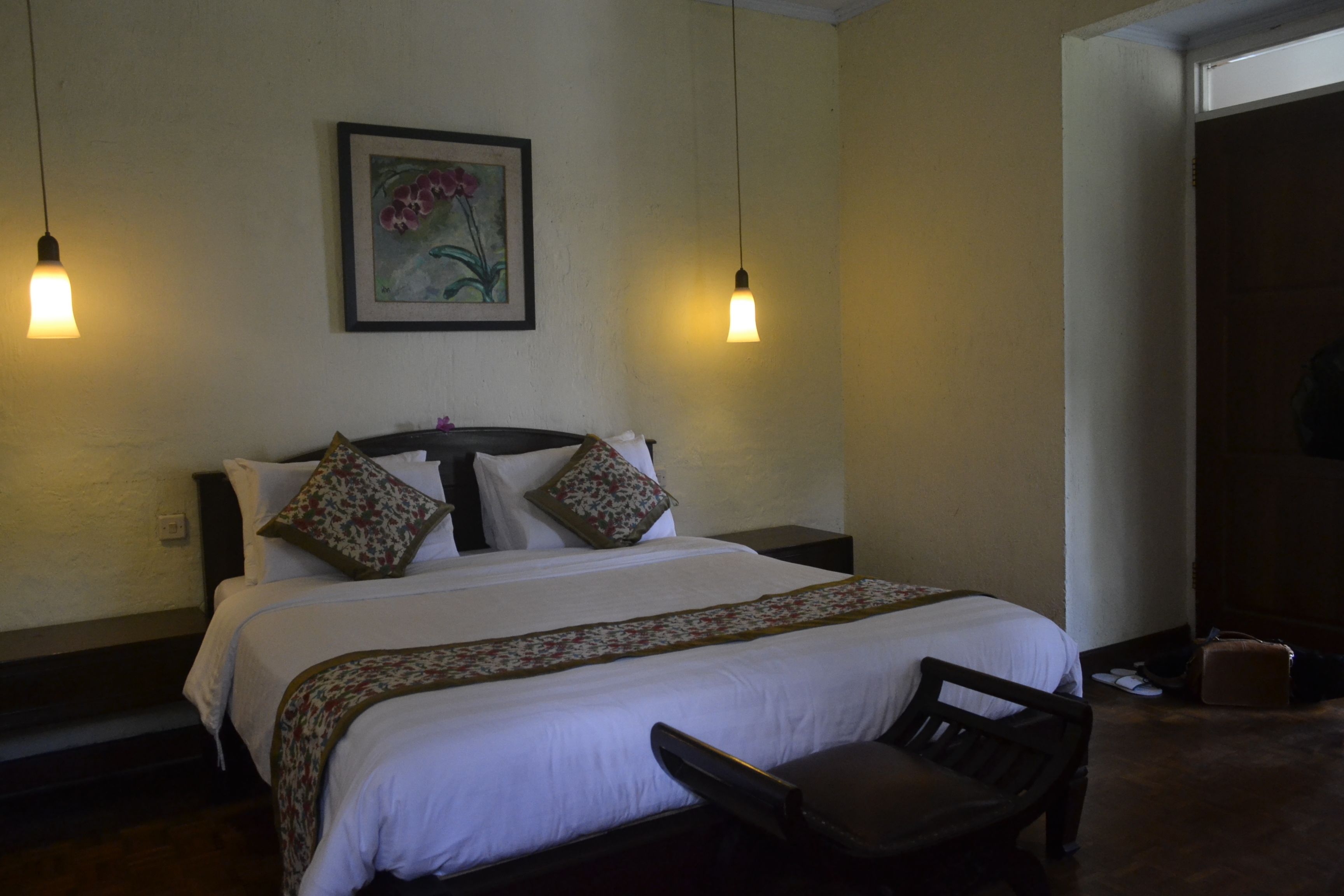 Een net opgemaakt bed met een bloemenbedekking, twee bedlampen en een lijst met een bloemistisch schilderij aan de muur.