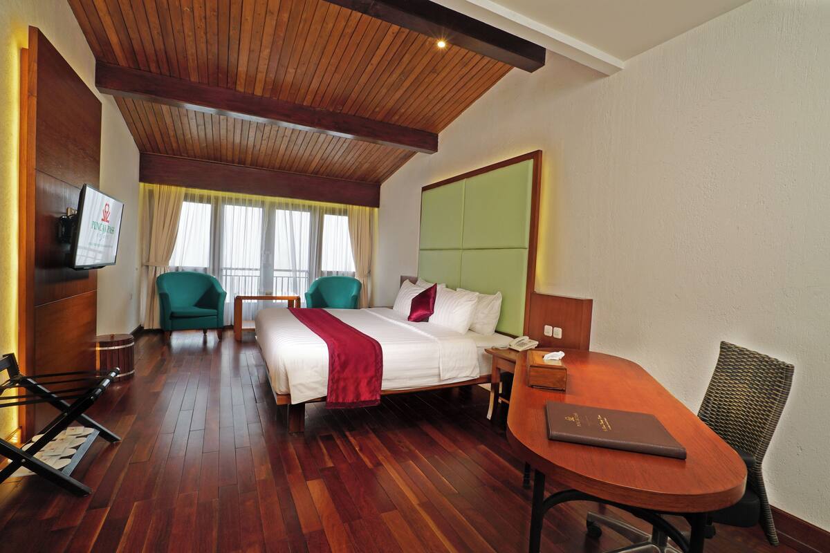 A hotel room with a wooden ceiling, a bed with white linens and a red throw, a desk with a laptop, and two chairs.