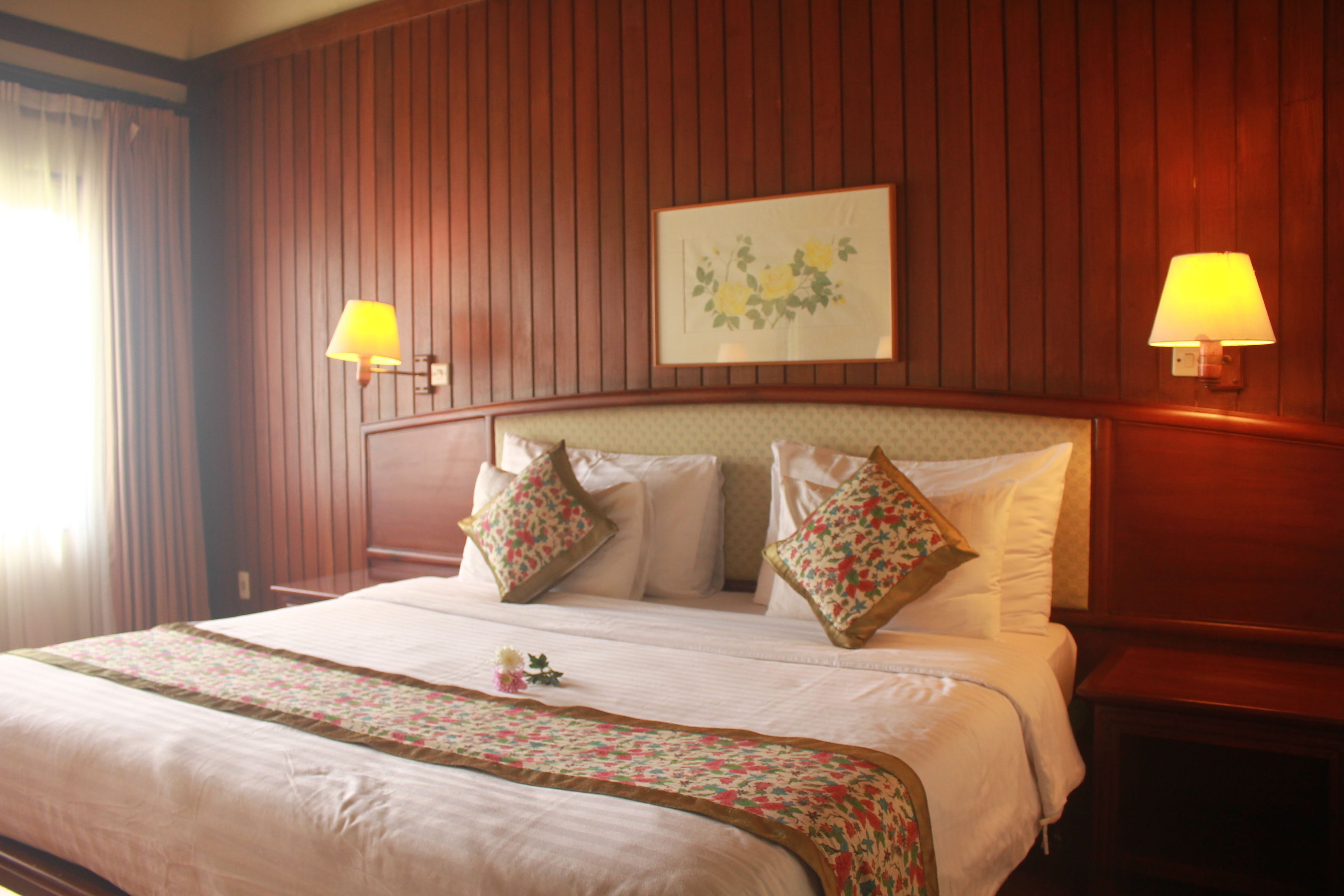 A hotel room with a large bed, two bedside lamps, a framed floral artwork, and a wooden headboard.