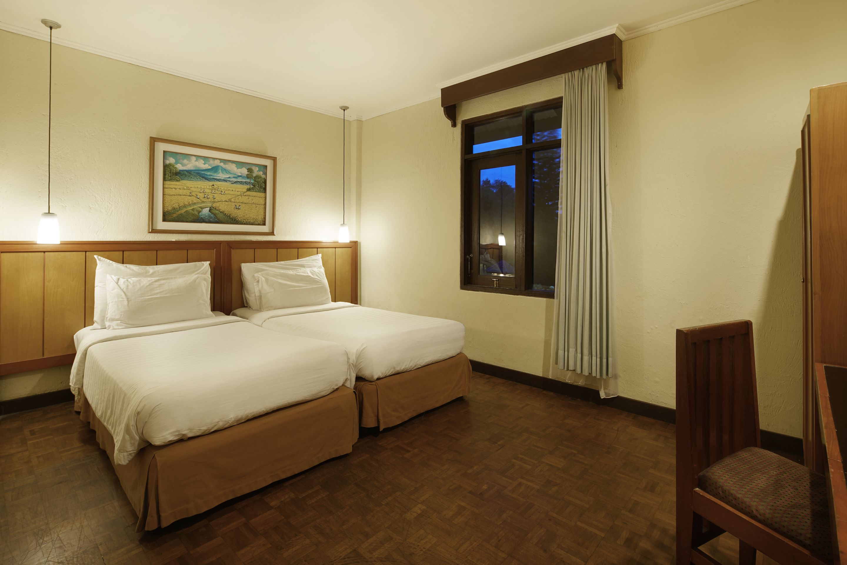 A hotel room with two beds, a wooden headboard, a painting on the wall, a window with curtains, and a chair.