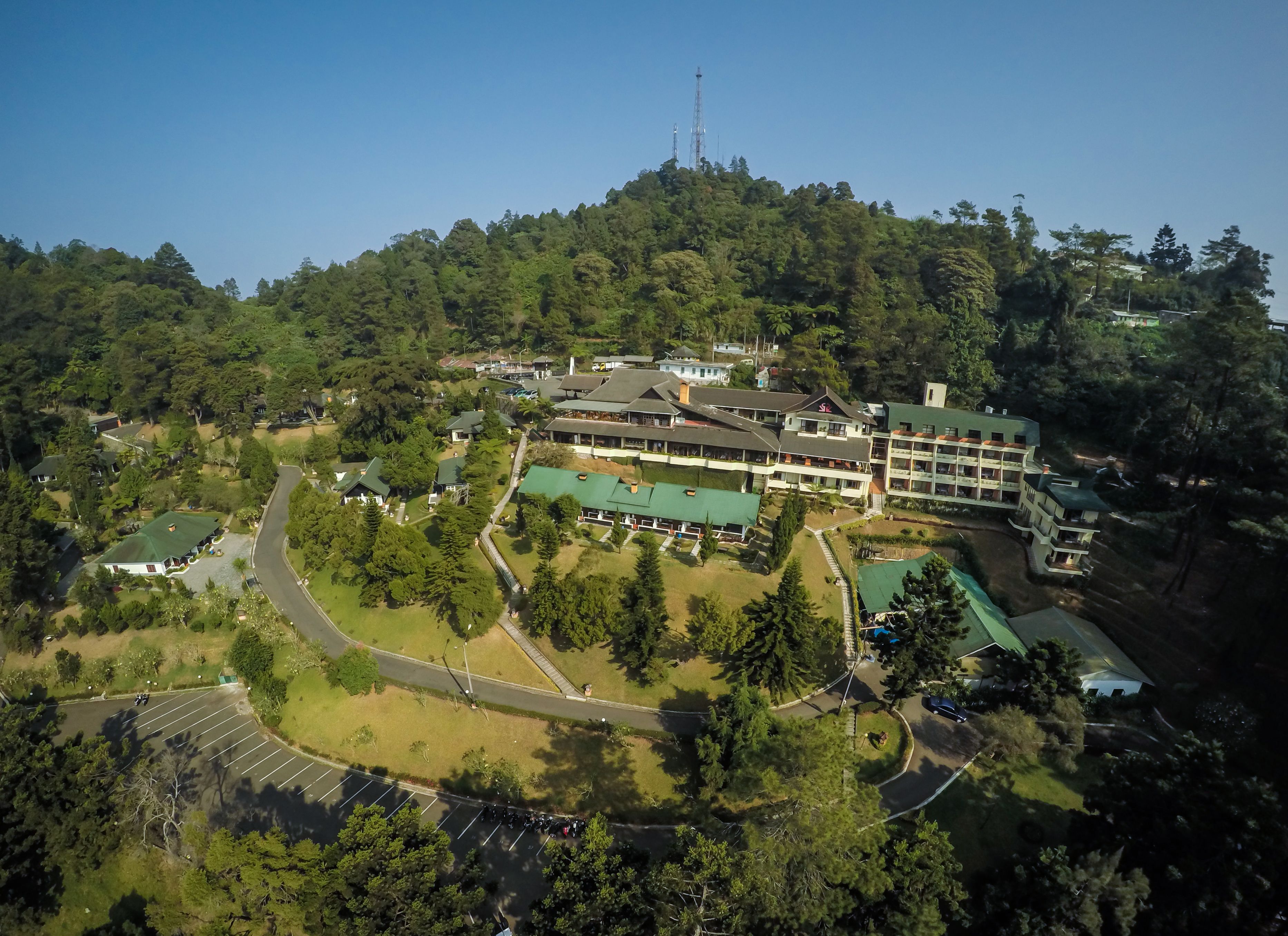 An aerial view of a hotel complex with multiple buildings, surrounded by dense forest. There are parking areas and a road visible.