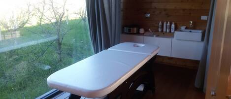 A wooden room with a massage table, a small sink, and a window overlooking a grassy area.