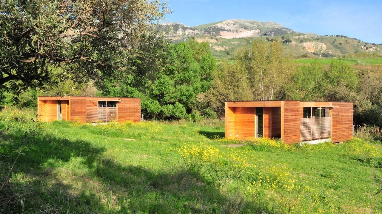 Two wooden cabins in a grassy field with trees and hills in the background.