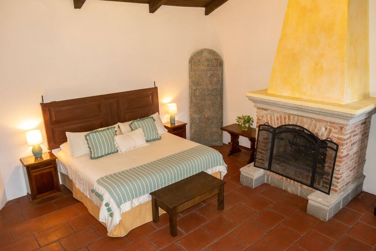 Un dormitorio con cama, mesitas de noche, un banco, chimenea y un elemento decorativo en la pared.