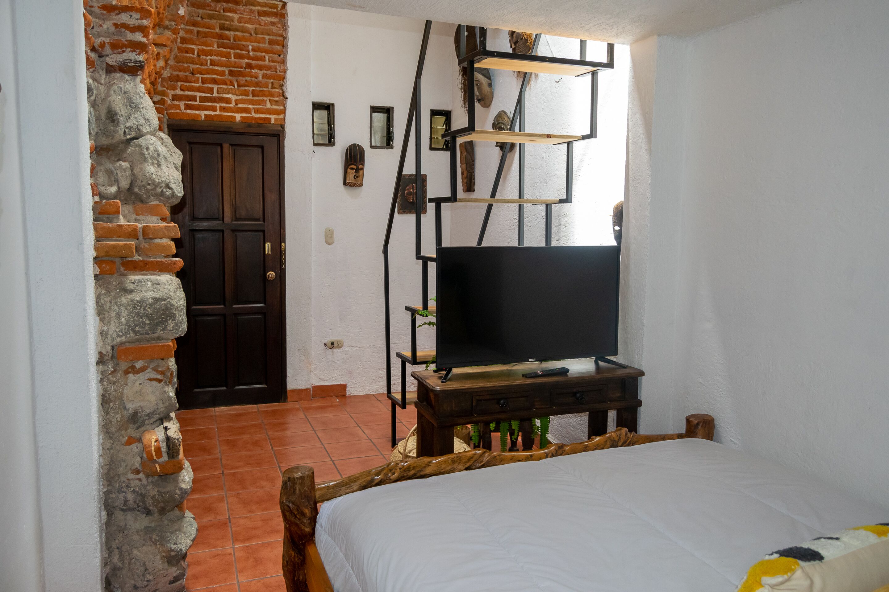Una habitación con una cama, un televisor sobre un soporte de madera, una pared de ladrillo y una puerta.