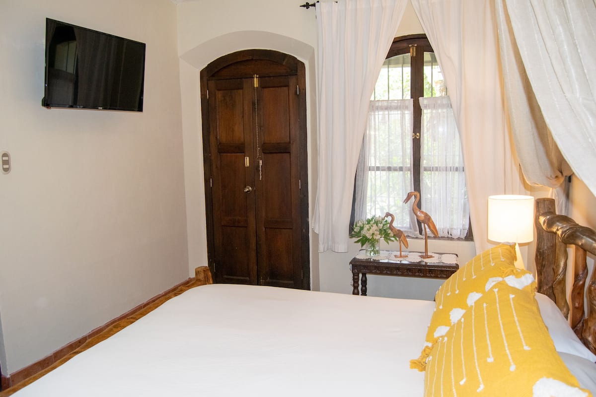 Un dormitorio con cama, una puerta de madera, una ventana con cortinas, una mesa con lámpara y un televisor montado en la pared.
