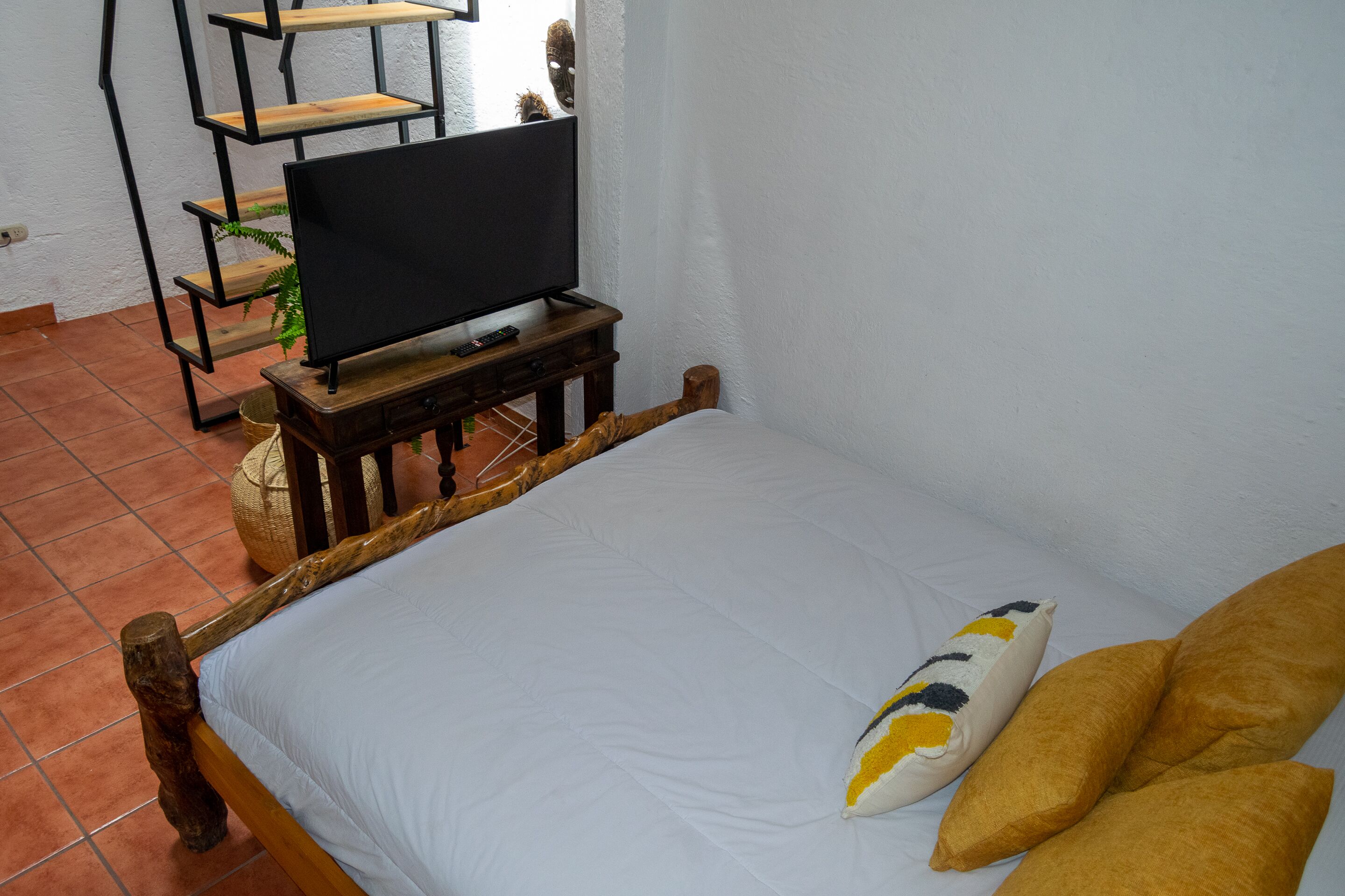 Una cama con cabecera de madera, un televisor sobre un mueble de madera y una pequeña estantería con una planta.