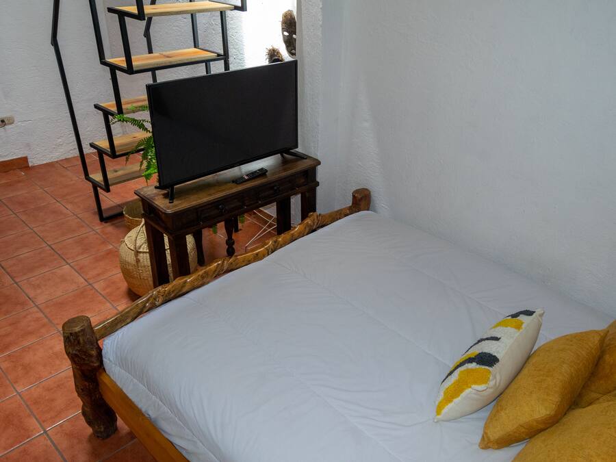 Ein Bett mit Holz-Kopfteil, ein Fernseher auf einem Holzboden und ein kleines Regal mit einer Pflanze.