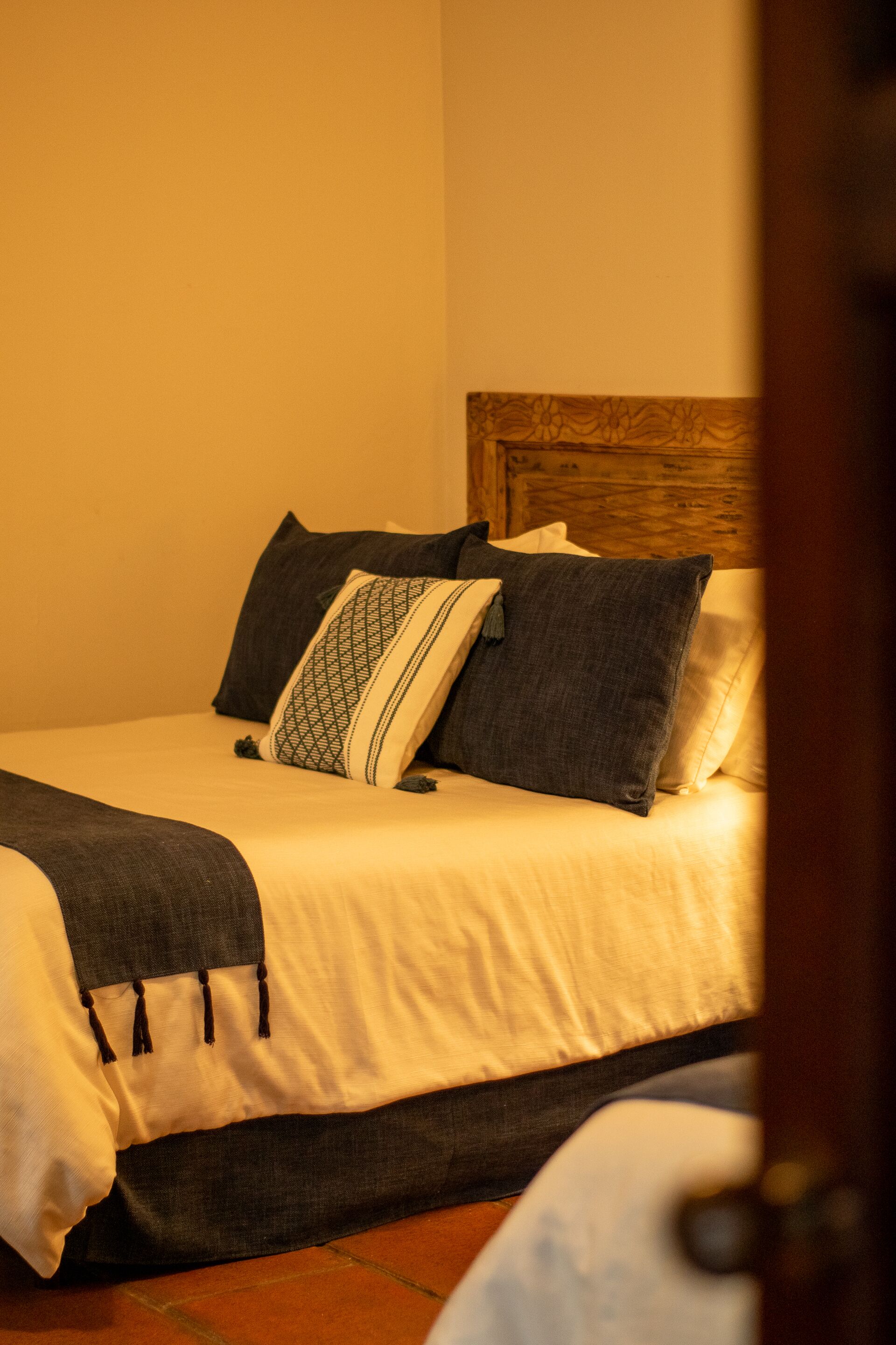 Una cama bien hecha con tablero de madera, ropa de cama blanca y oscura, y una almohada estampada.