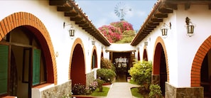 A courtyard with arched doorways, a windmill in the background, and a pathway leading to a gate. - Majoro (Canaria)