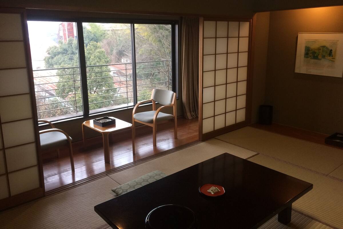 A traditional Japanese room with tatami flooring, a low table, two chairs, a sliding shoji screen, and a view of trees and buildings outside.