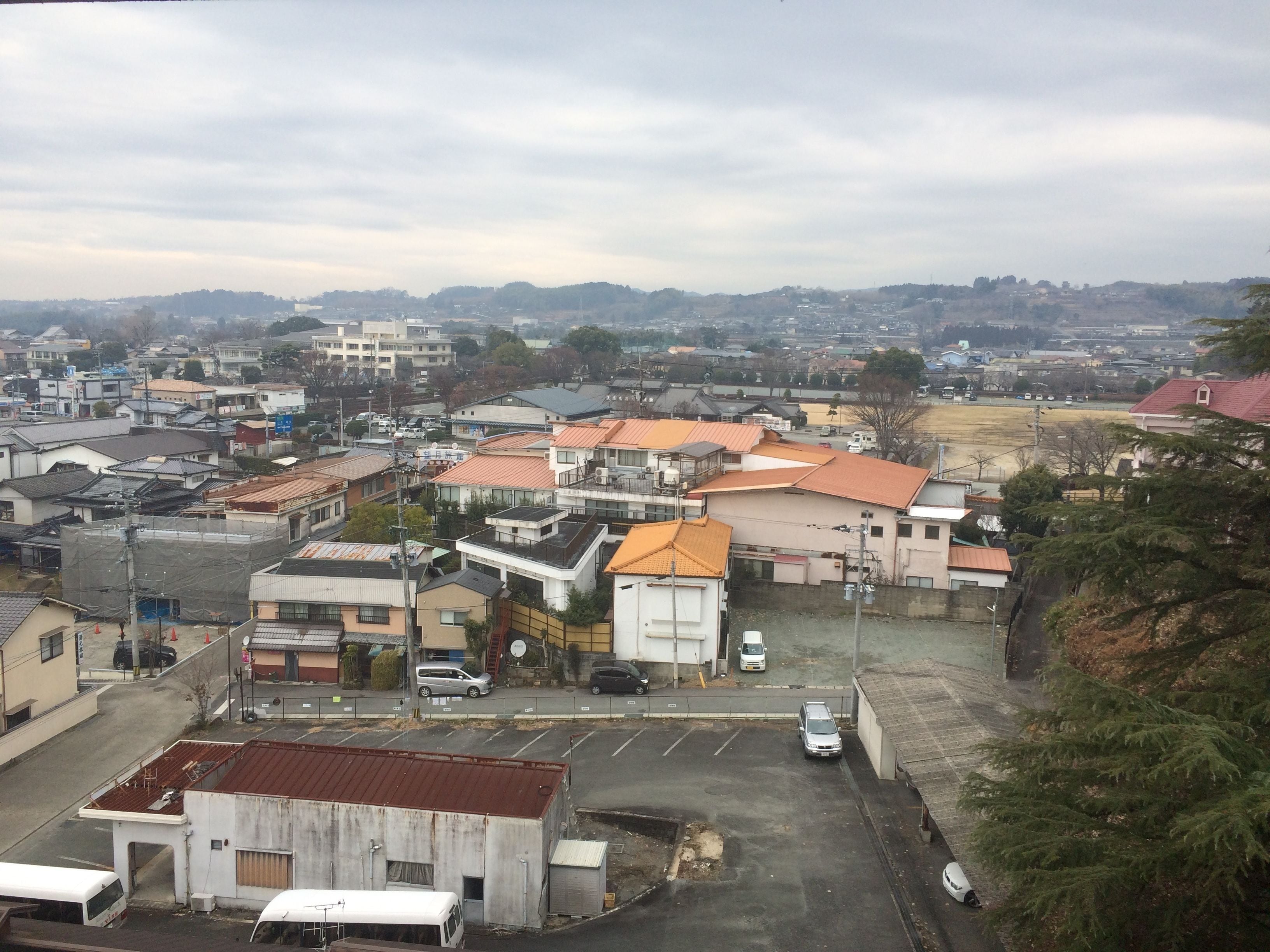 A residential area with various buildings, including houses and a small commercial structure. There is a parking lot with several cars and a bus.