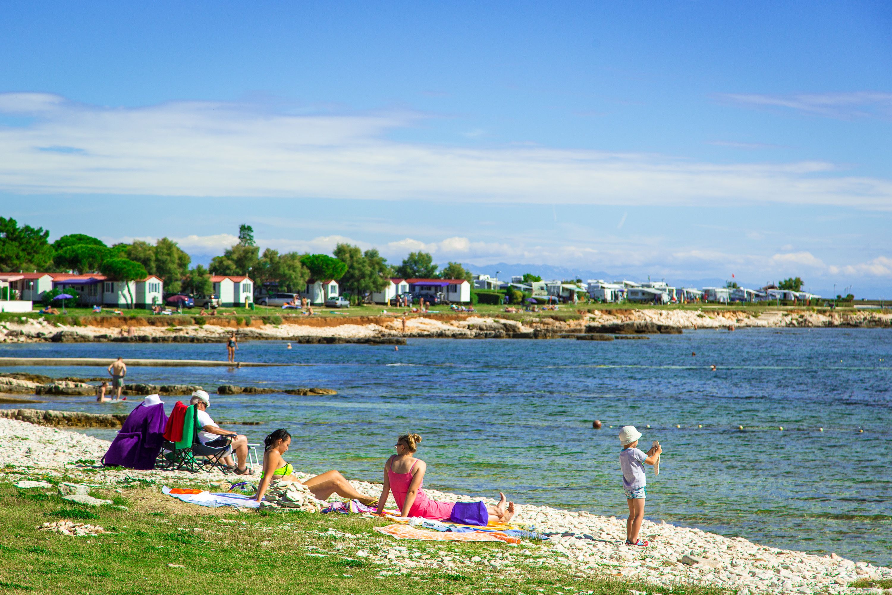 Ein Strandbild mit Menschen, die auf Handtüchern und Liegestühlen entspannen, einem Gewässer und einer Reihe von Häusern im Hintergrund.