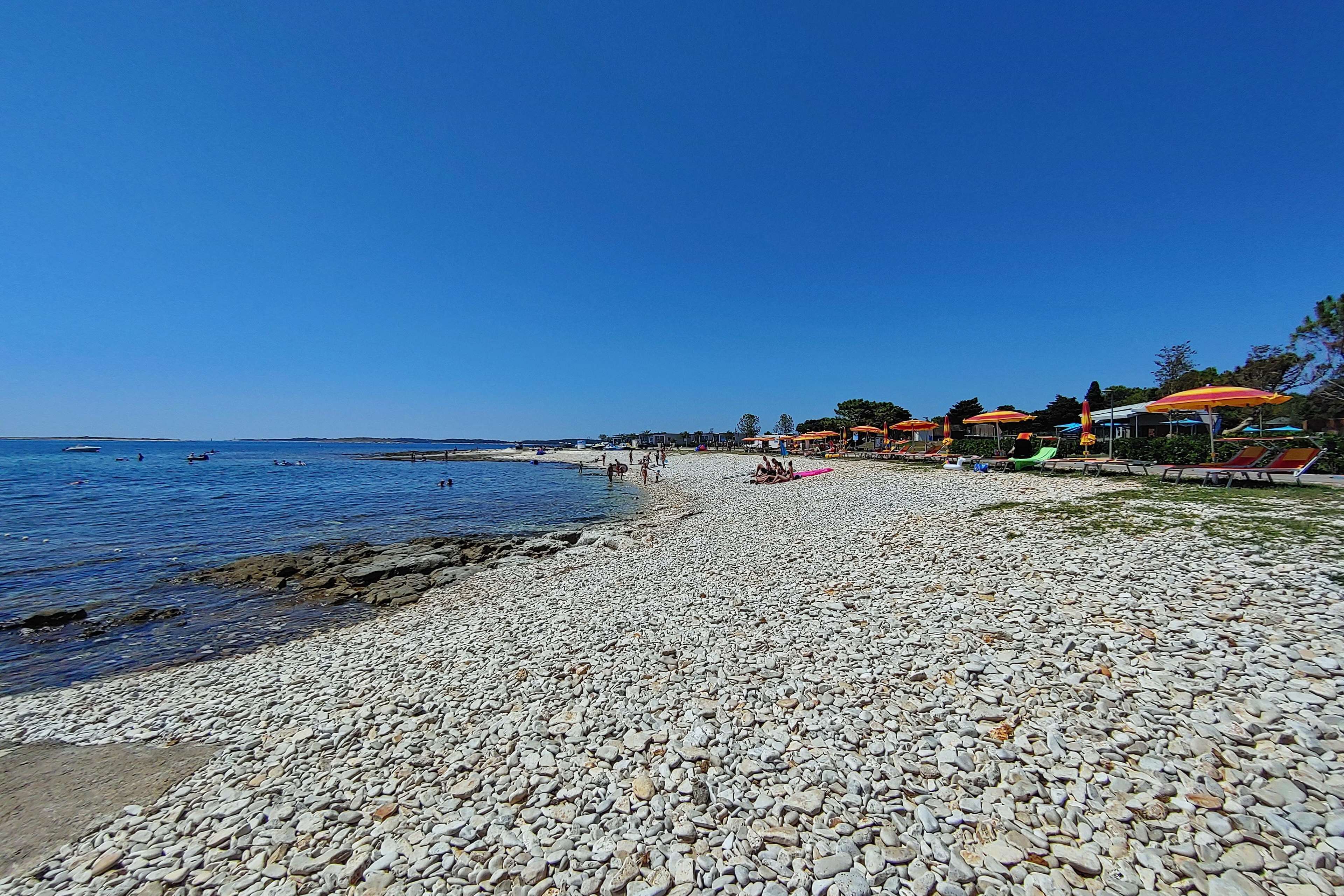 A pebble beach with umbrellas and lounge chairs, a clear blue sky, and the sea in the background.