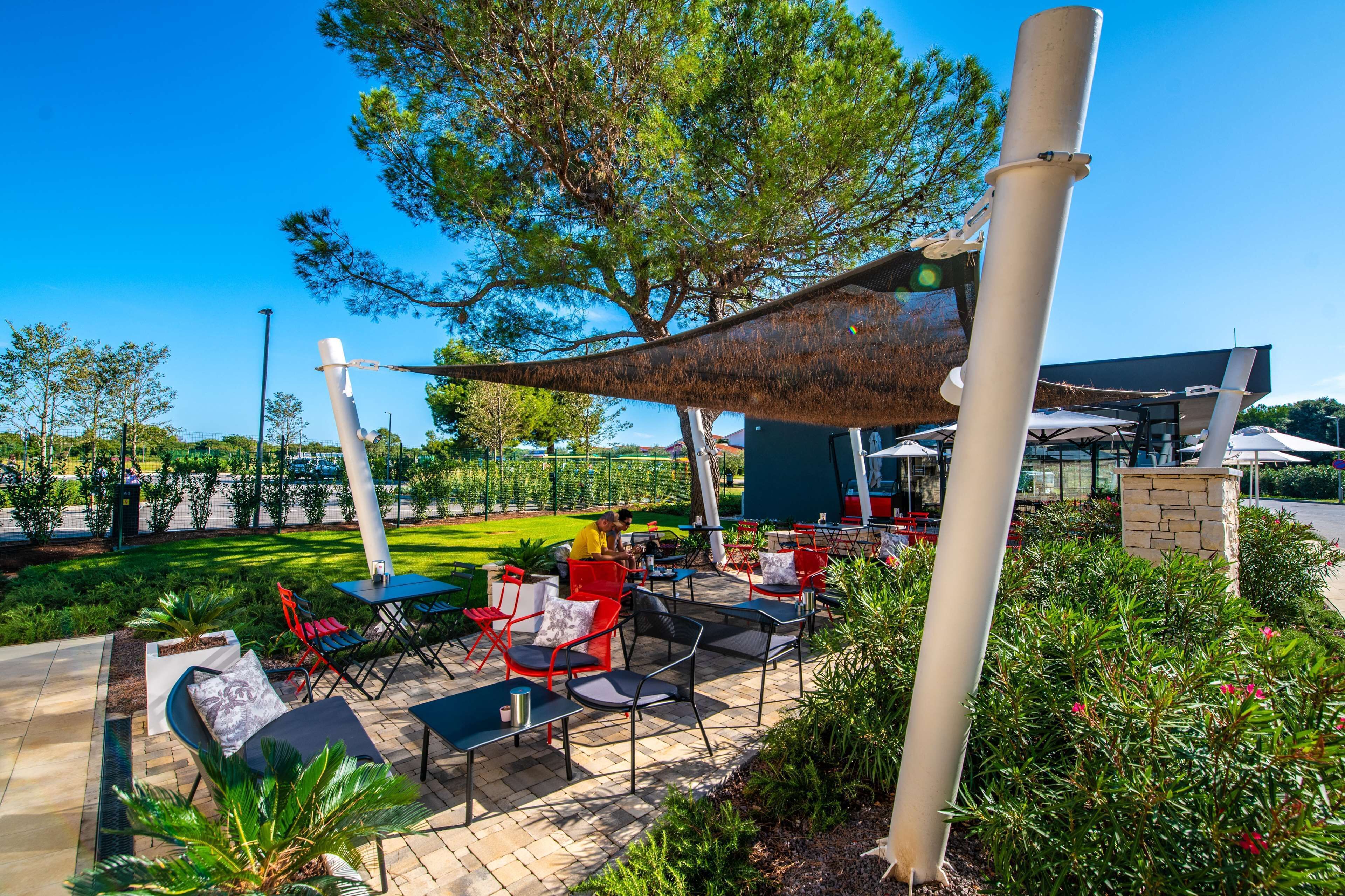 An outdoor patio area with tables and chairs, surrounded by greenery and a large tree.