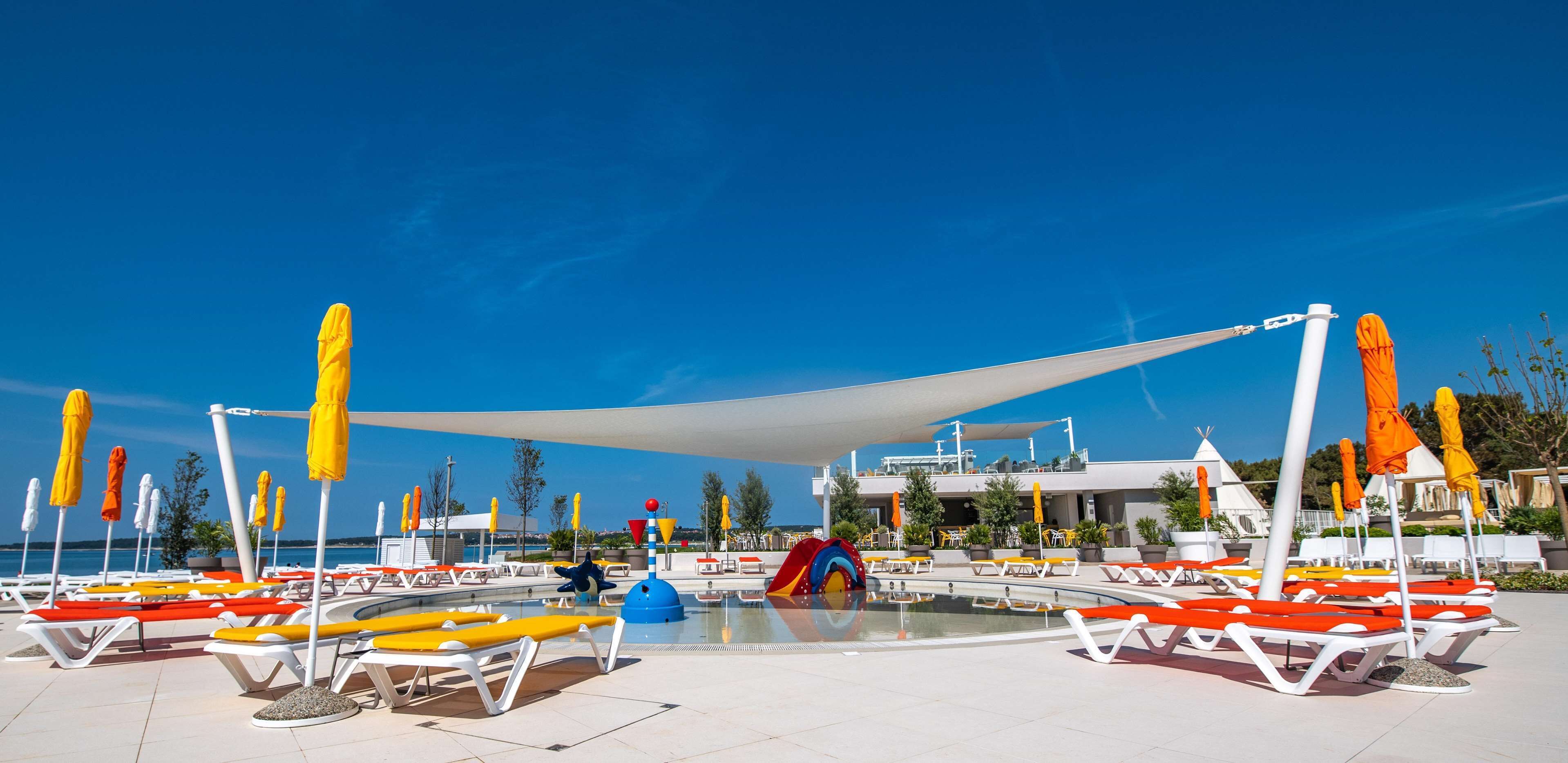 Zona junto a la piscina con tumbonas, sombrillas y un parque infantil.