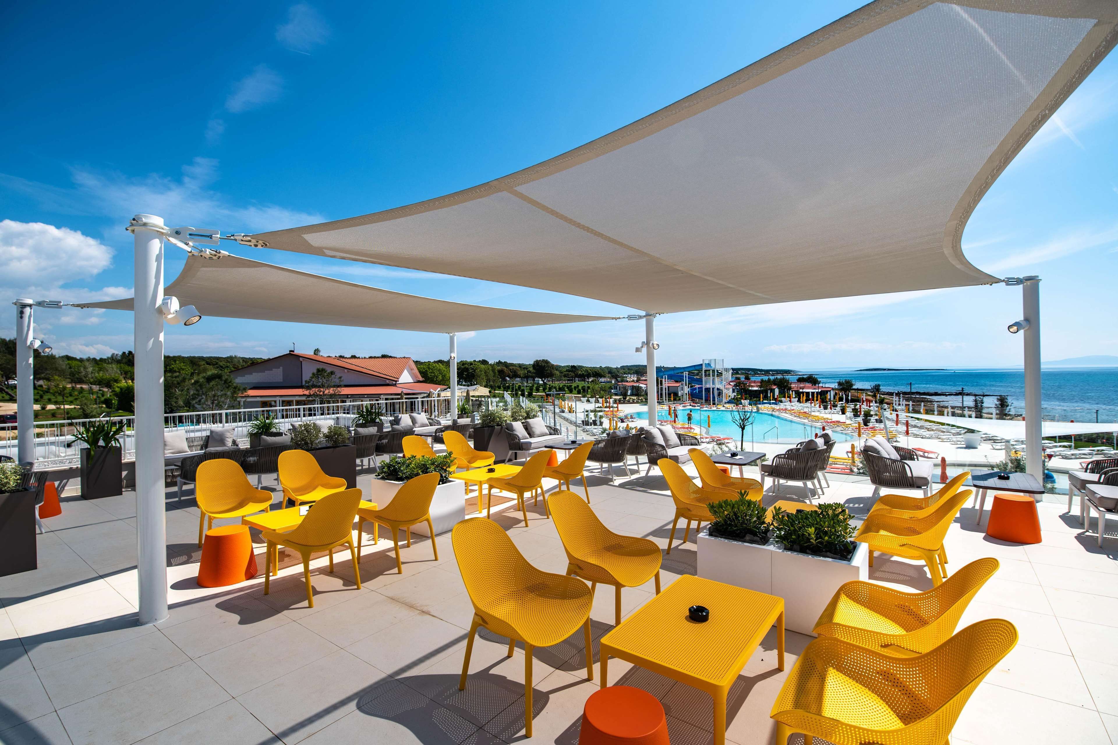A rooftop terrace with yellow chairs and tables, a pool, and a view of the sea.