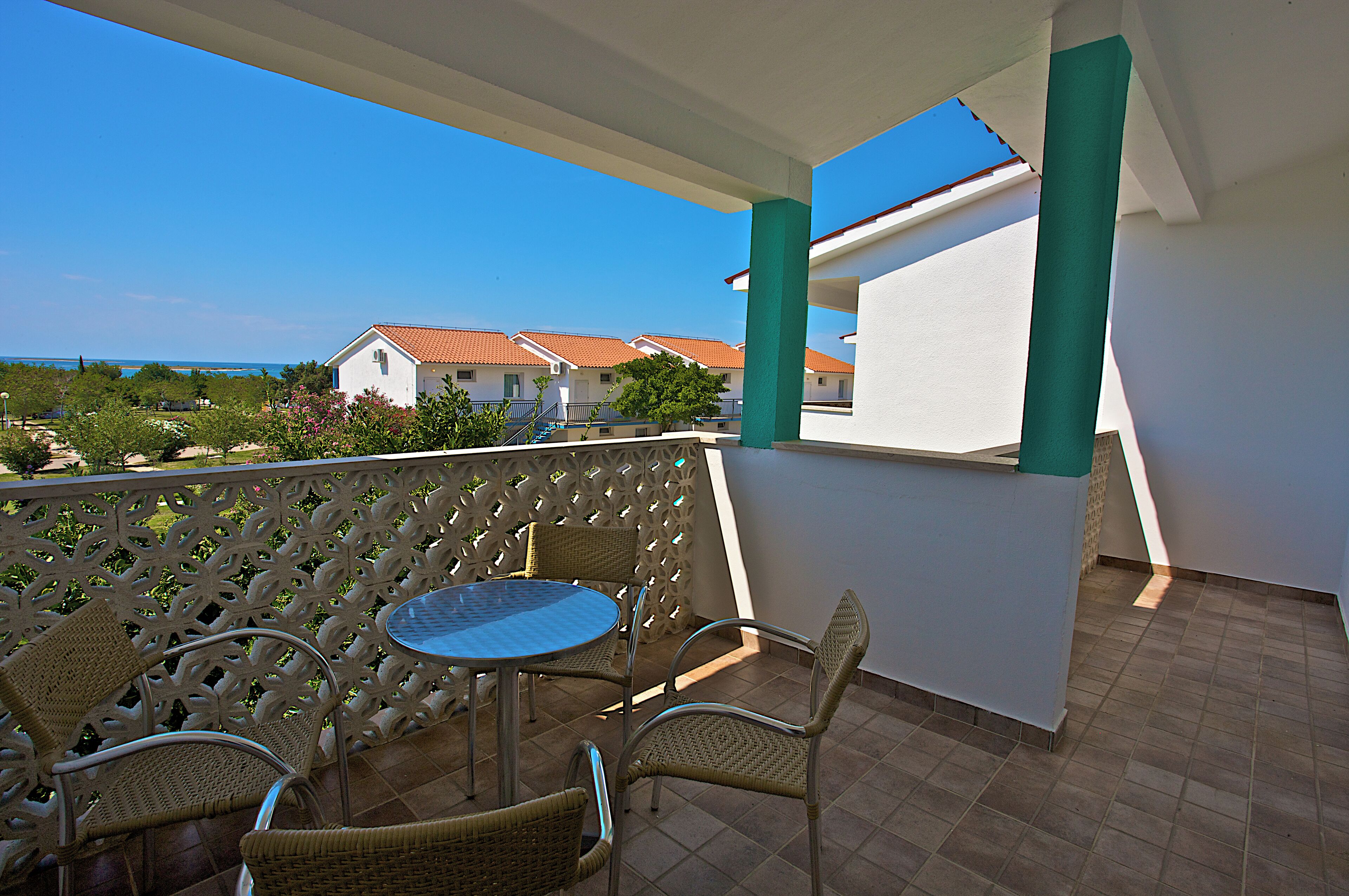 A balcony with wicker chairs and a table, overlooking a coastal area with buildings and greenery.