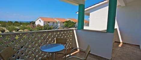 A balcony with wicker chairs and a table, overlooking a coastal area with buildings and greenery.