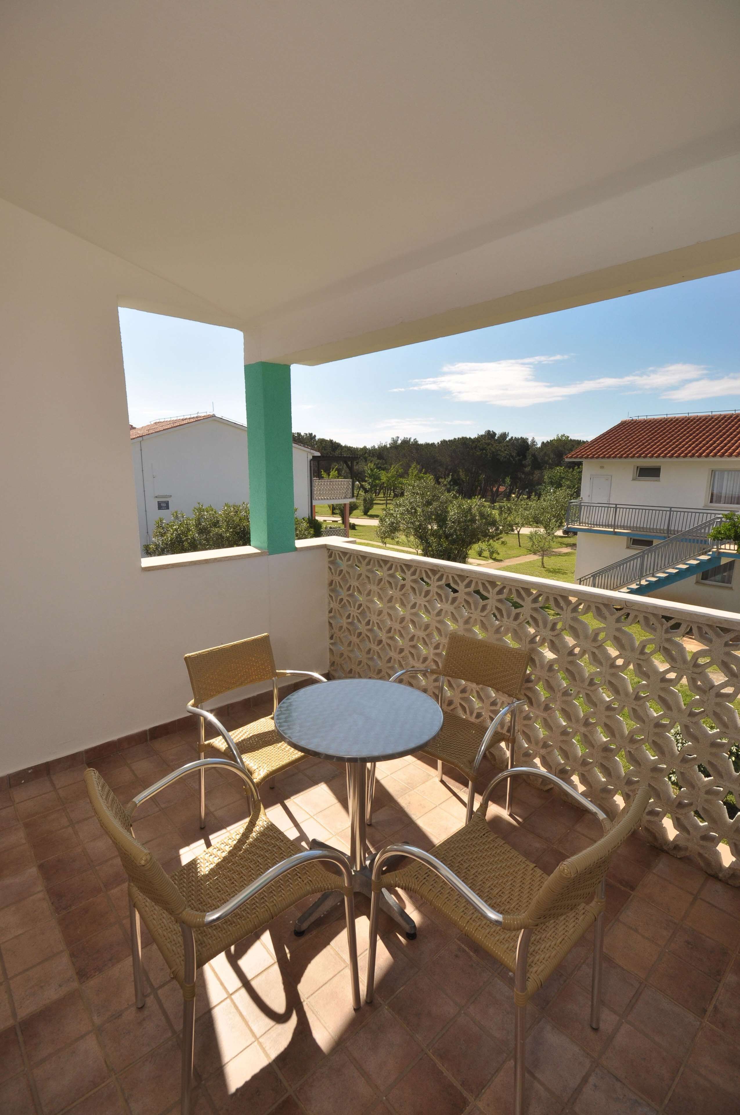 A balcony with a round table and four wicker chairs, a view of a residential area, and a green pillar.
