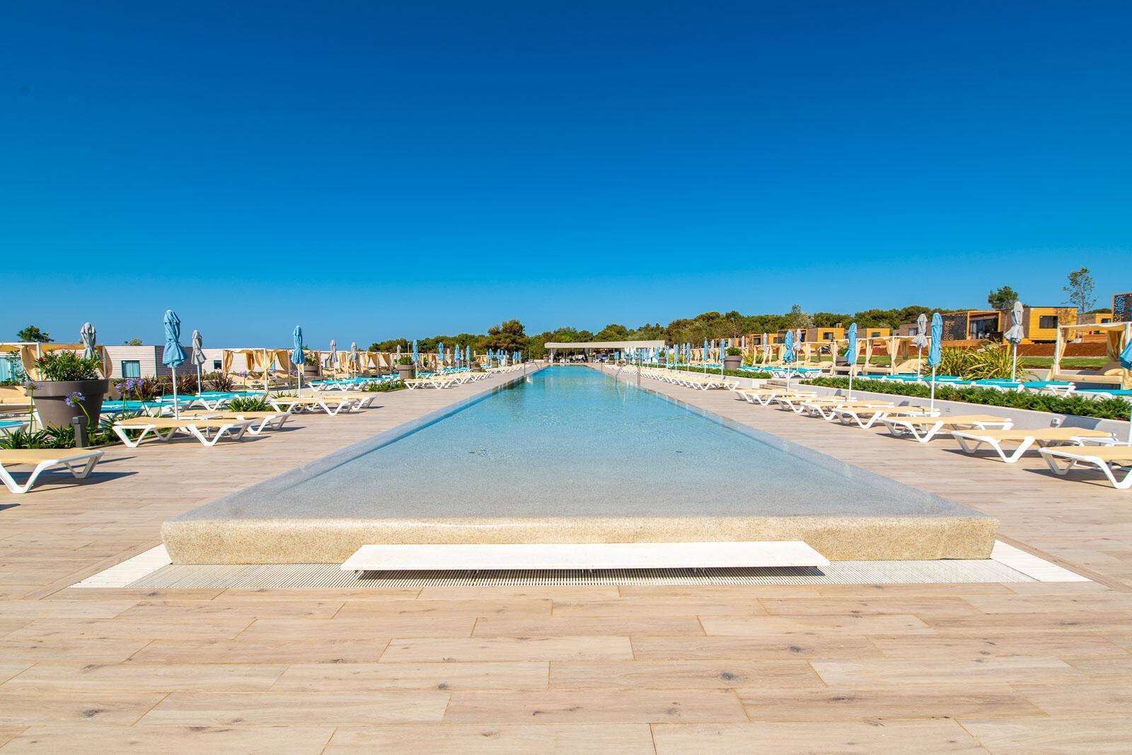 A long swimming pool with lounge chairs and umbrellas on both sides, surrounded by buildings.