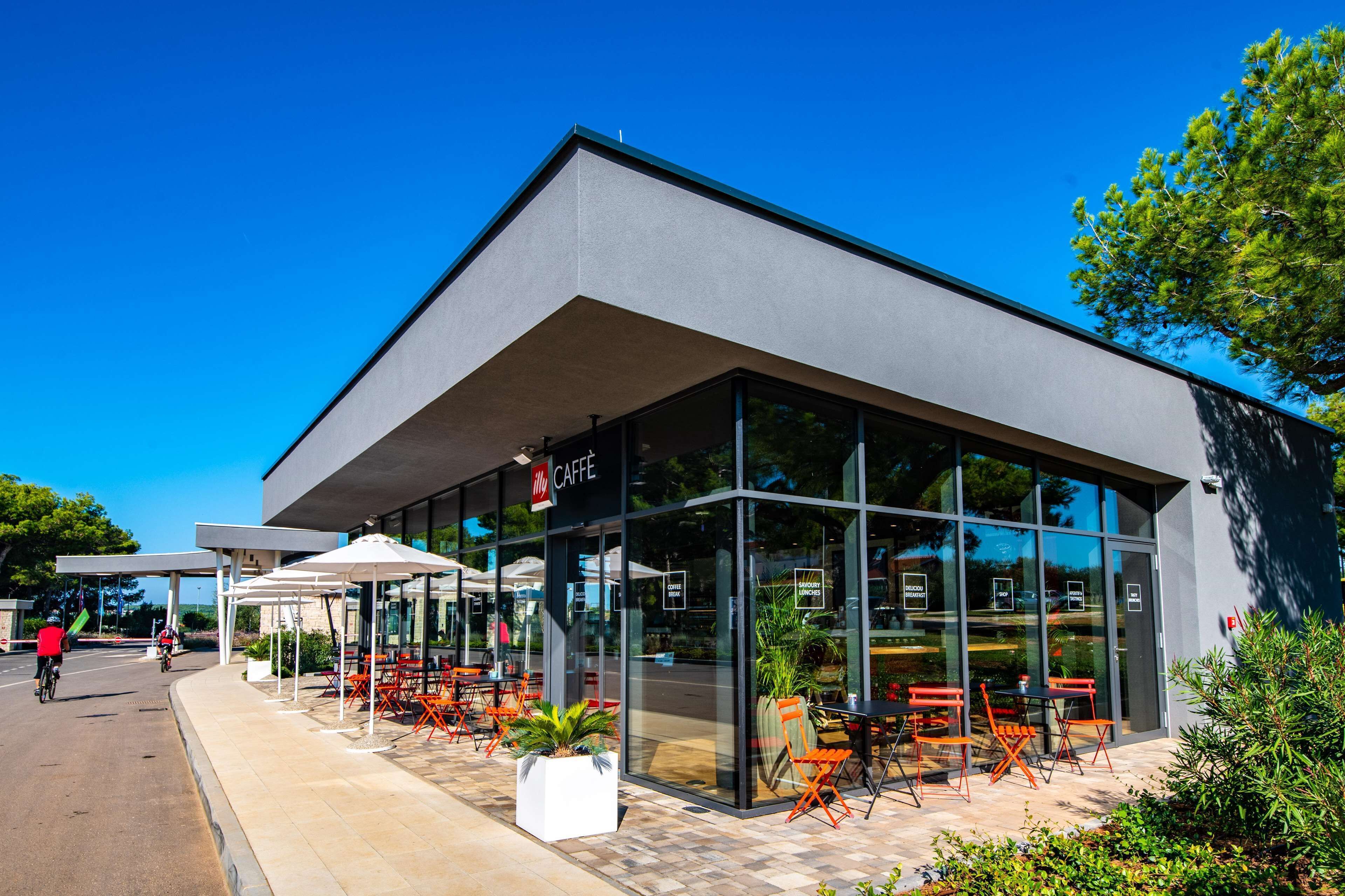 Ein modernes Gebäude mit einem Café im Erdgeschoss, Außenbestuhlung und blauem Himmel.