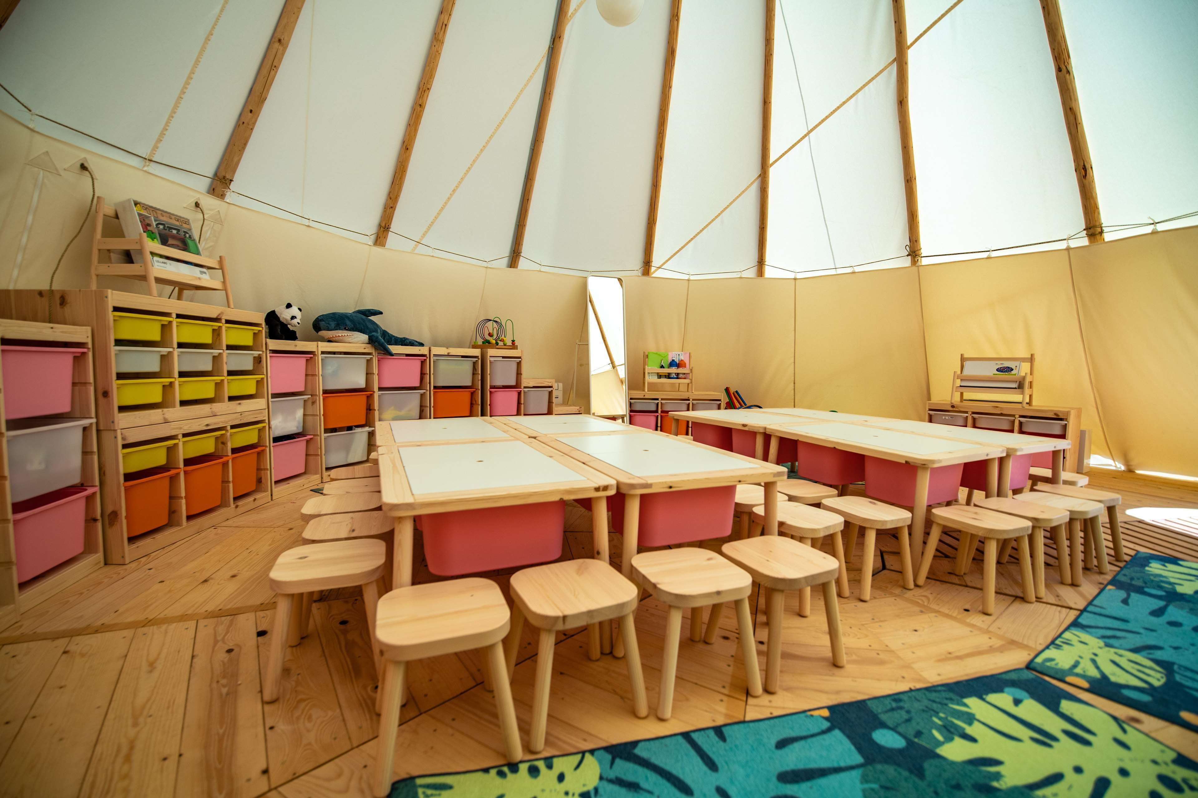 A classroom with wooden tables and chairs, colorful storage bins, and a large tent structure.