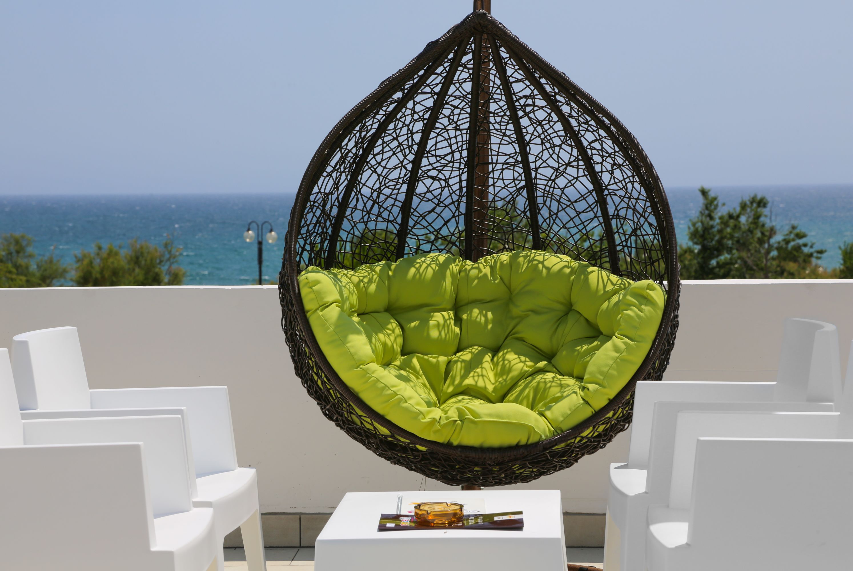 A hanging chair with a green cushion, white chairs, and a small table with a magazine on a balcony with a view of the sea.
