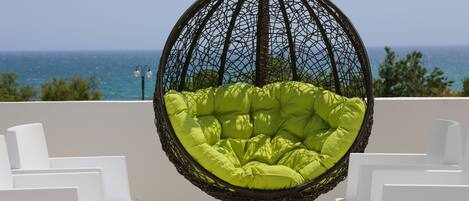 A hanging chair with a green cushion, white chairs, and a small table with a magazine on a balcony with a view of the sea.