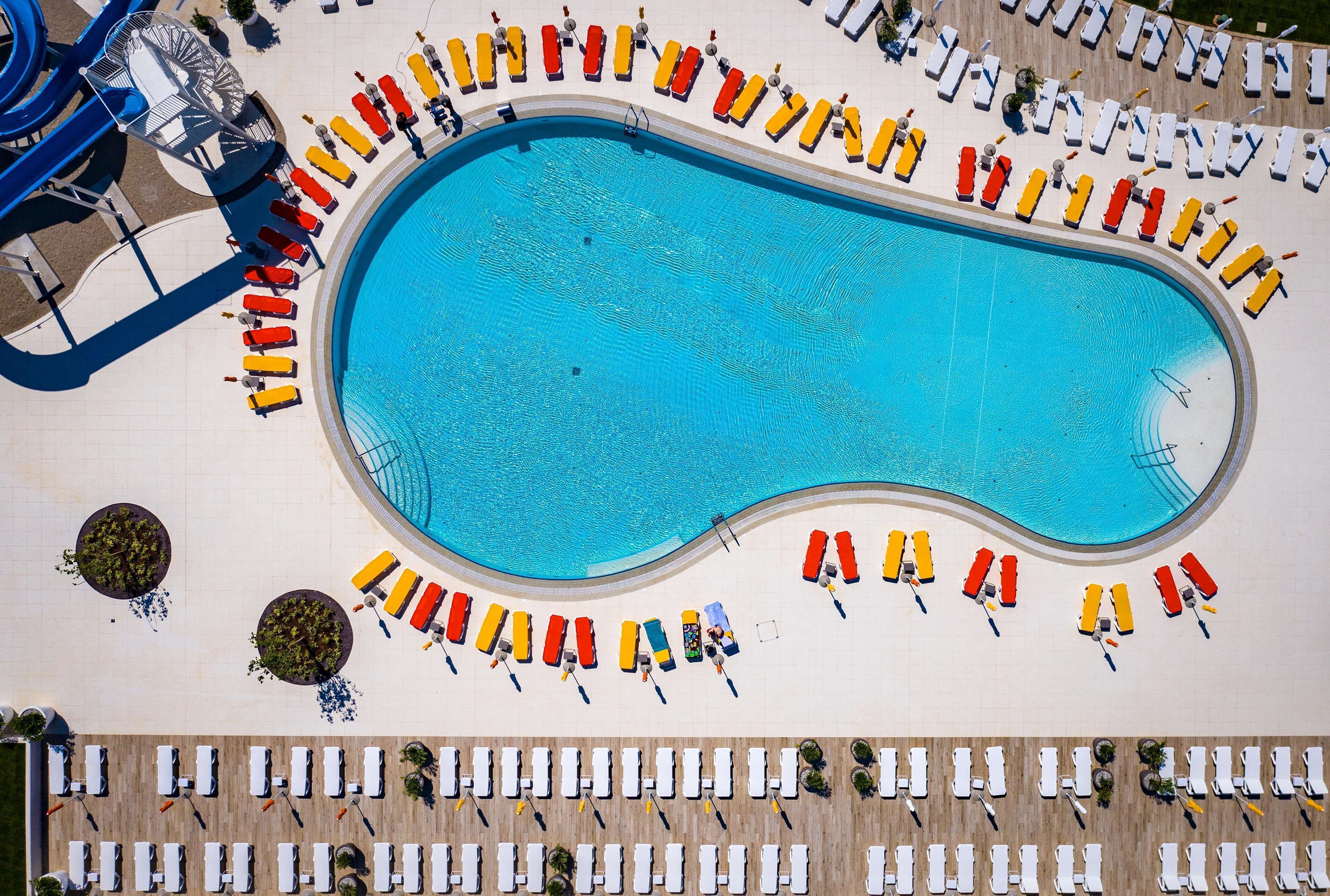 A large swimming pool surrounded by rows of sun loungers and chairs, with a water slide and a covered area in the background.