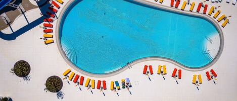 A large swimming pool surrounded by rows of sun loungers and chairs, with a water slide and a covered area in the background.