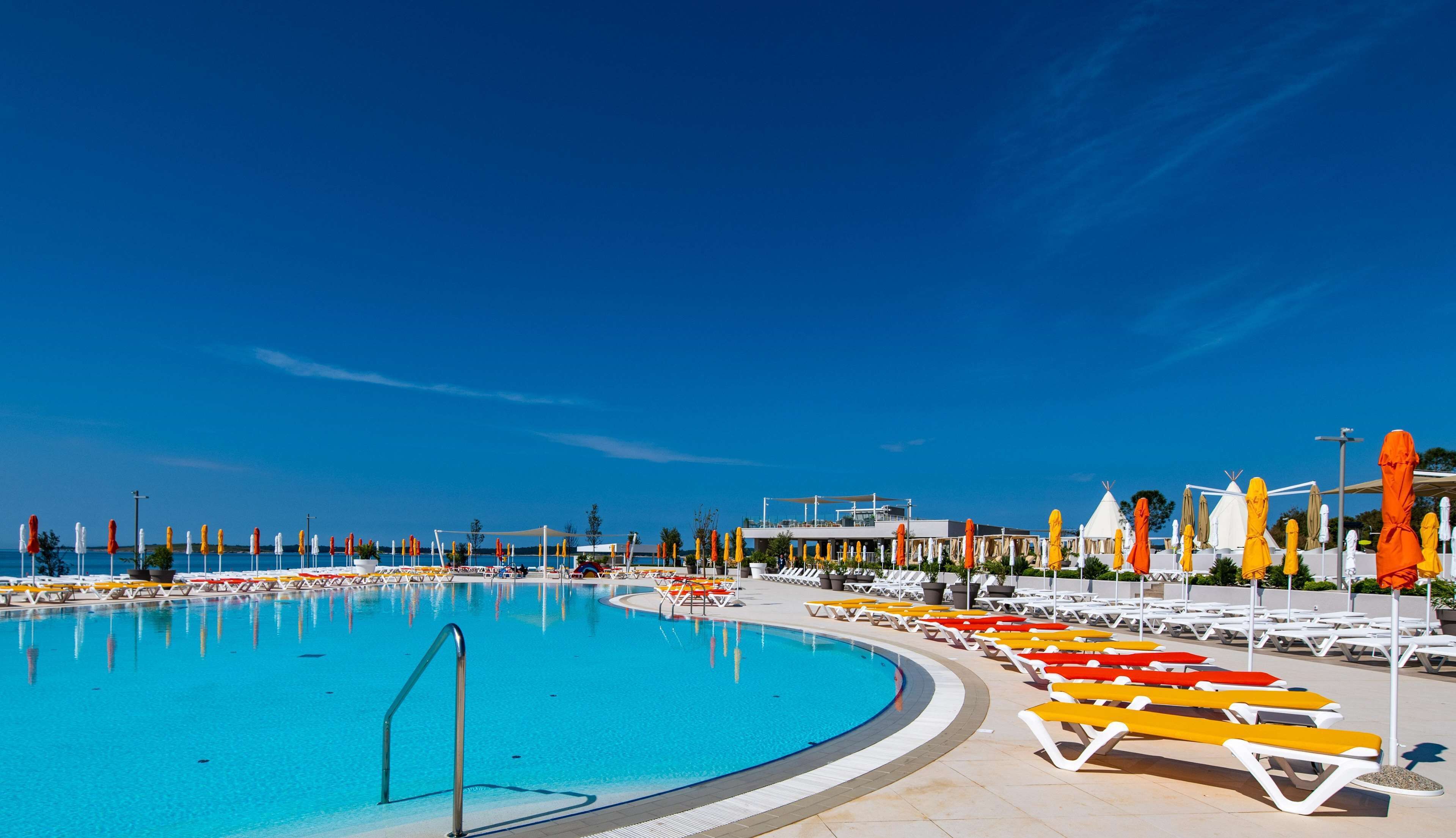 A large outdoor swimming pool with multiple sun loungers and umbrellas, surrounded by buildings and a clear blue sky.
