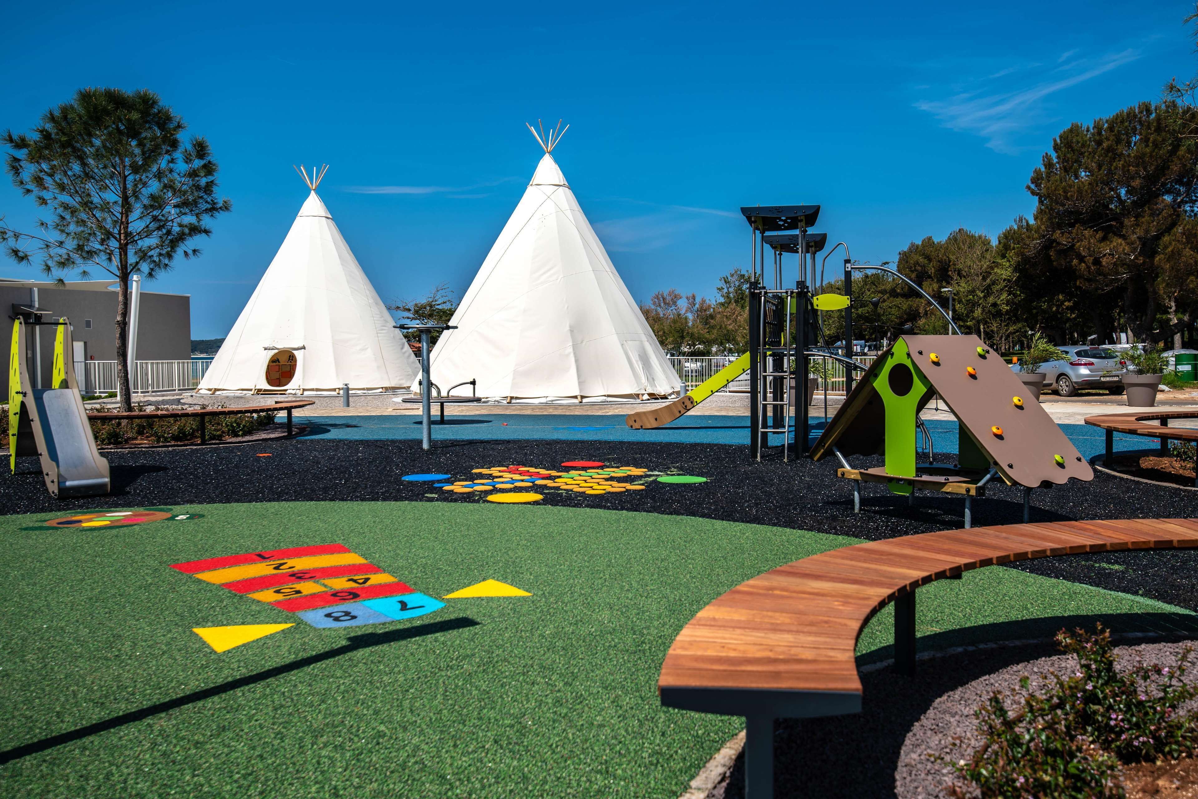 Ein Spielplatz mit zwei weißen Teepeezeltürmen, einer Rutsche und einer Kletterkonstruktion.