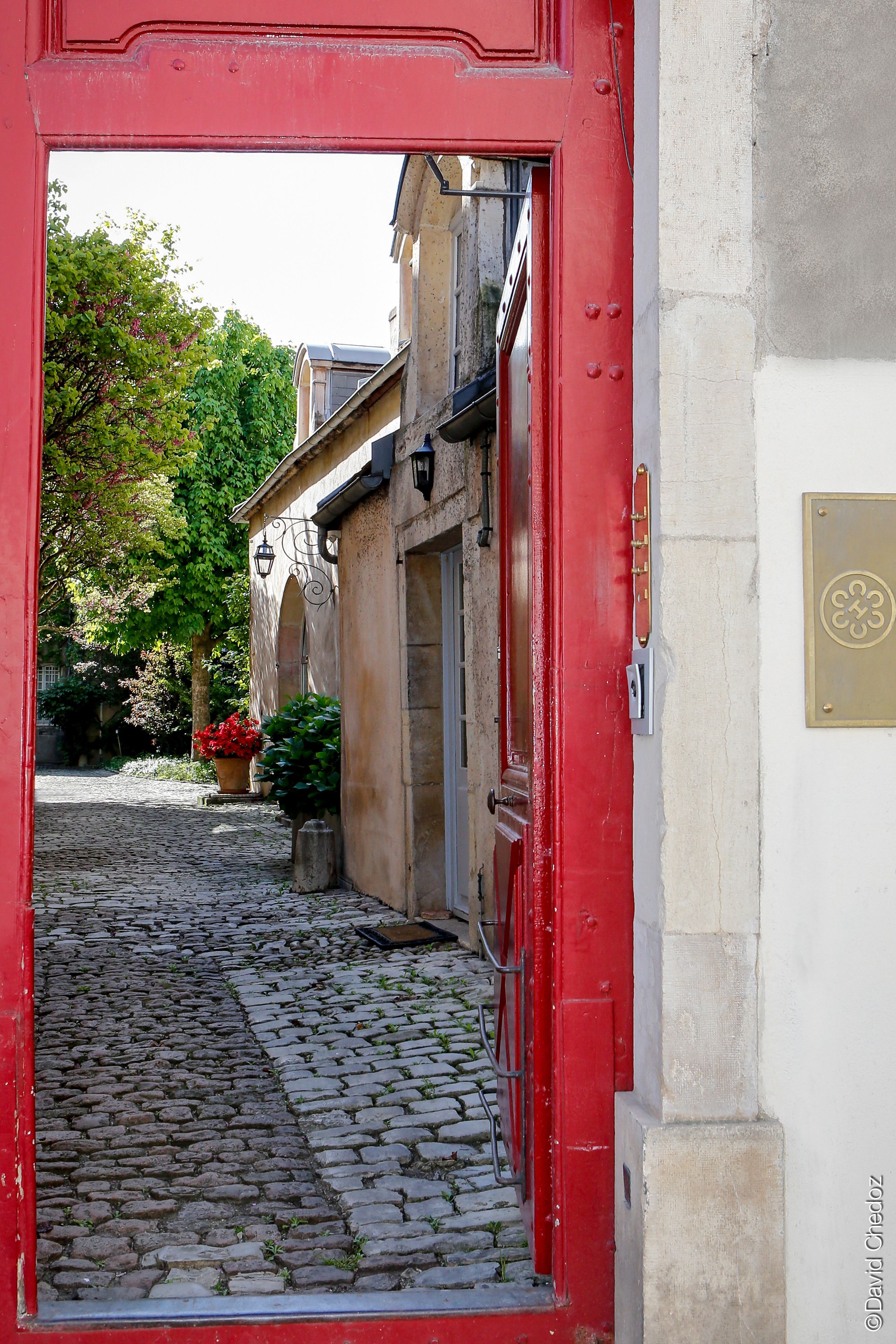 A cobblestone alleyway with a red door leading to a building with a stone facade and a brass plaque.