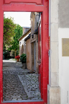 Un callejón adoquinado con una puerta roja que da a un edificio de fachada de piedra y placa de latón.