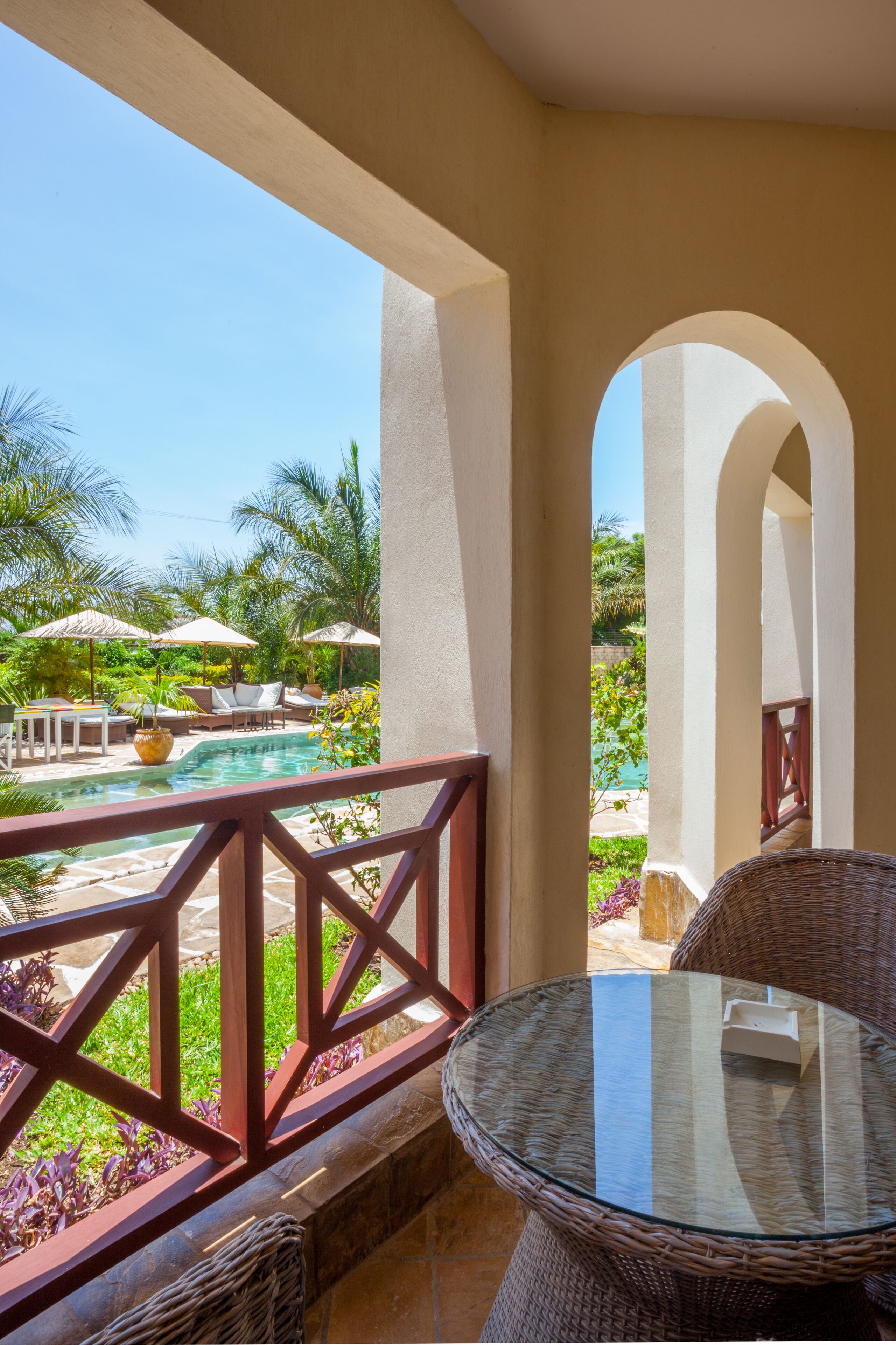 A balcony with a glass table, wicker chairs, and a view of a pool and palm trees.