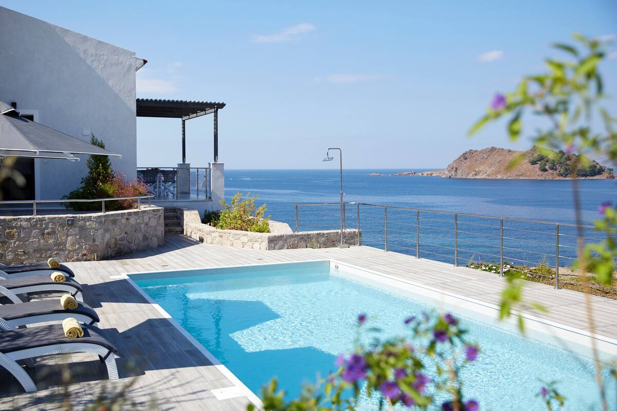 A pool area with lounge chairs, a stone wall, and a view of the sea.