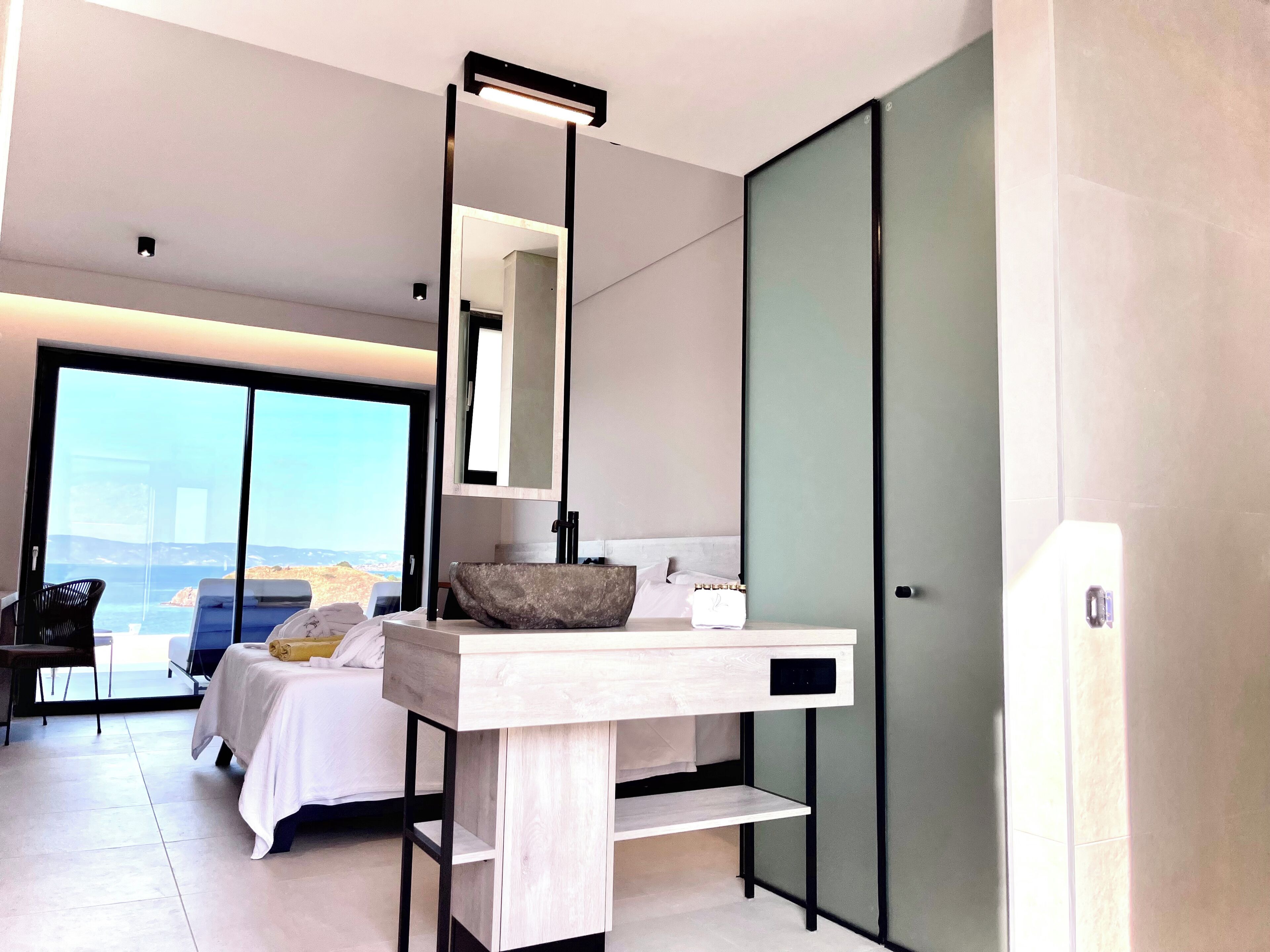 A modern hotel room with a large bed, a bedside table with a mirror, a bathroom with a large mirror, and a view of the sea.